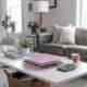 A cozy living room with a white coffee table, holding a pink binder, calculator, plant, and coffee cup. Neutral-toned sofa, cushions, and a lamp in the background.