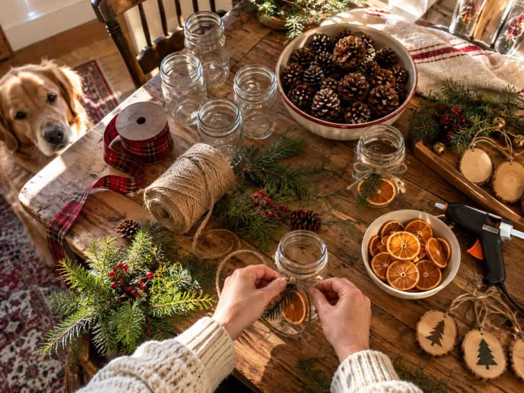 30 Cheap DIY Christmas Decorations That Look Like a Million Bucks 2 Person creating DIY Christmas decorations with pinecones and jars at home.